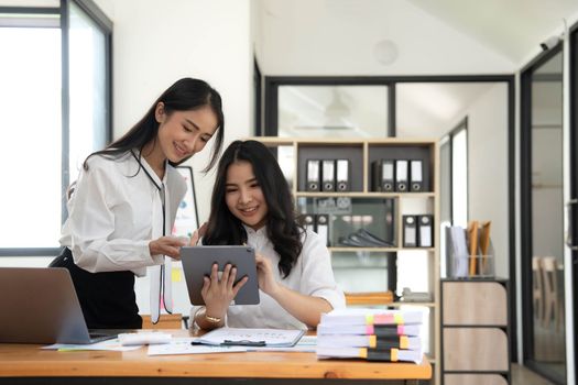 Two young Asian businesswomen show joyful expression of success at work smiling happily with a laptop computer in a modern office..