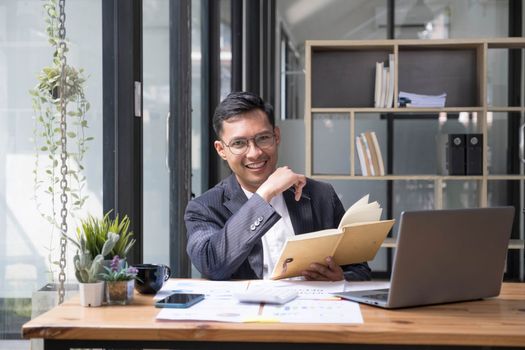 Young Asian businessman in suit and holding hot coffee cup and looking on laptop computer in office. Man in suit using laptop in well-lit workplace...