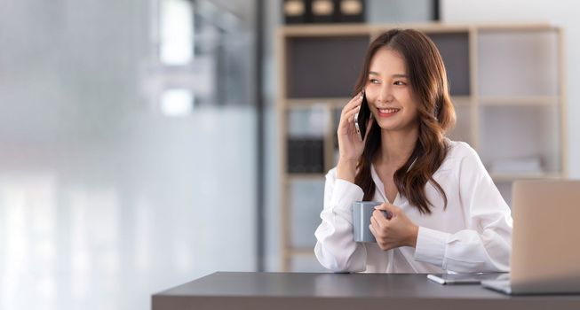 Young Asian businesswoman sit with her laptop computer happily talking on the phone with her customer explaining the detail in the office...