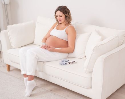 Pregnant woman sits on a white sofa and holds a smartphone. Fetal ultrasonic screening