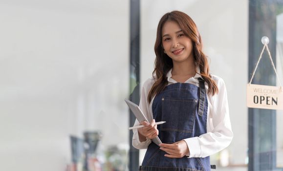 Beautiful asian young barista woman in apron holding tablet and standing in front of the door of cafe with open sign board. Business owner startup concept...