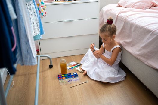 Caucasian child girl is painting watercolor paint on small ceramic figure with fun. Kids arts and crafts creative activity class at home. Little girl make ceramic decor sitting floor