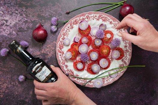 As,Belgium, May 10, 2022: tomato salad with purple onion rings dressed with Ponti balsamic dressing and olive oil with feta cheese and olives on a dark purple background, step by step recipe,flat lay