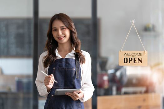 Beautiful asian young barista woman in apron holding tablet and standing in front of the door of cafe with open sign board. Business owner startup concept...