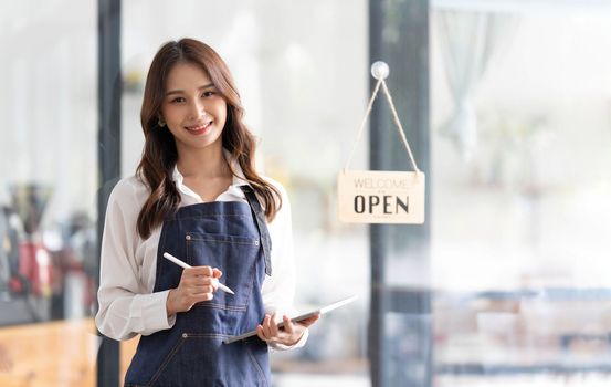 Beautiful asian young barista woman in apron holding tablet and standing in front of the door of cafe with open sign board. Business owner startup concept...