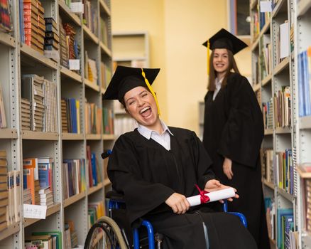 Happy young woman and woman in wheelchair in graduate gown with diploma in hands in library. Inclusive education