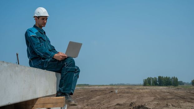 Caucasian male builder in hardhat sits on floor slabs and uses laptop at construction site