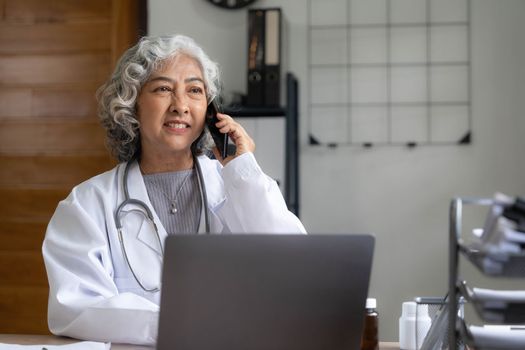 Senior asian female doctor using mobile phone talking while sitting in hospital office. happy woman medical worker having joyful conversation with husband in break time. nurse resting in clinic..