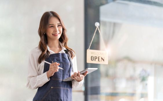 Beautiful asian young barista woman in apron holding tablet and standing in front of the door of cafe with open sign board. Business owner startup concept...