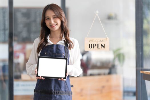 Beautiful asian young barista woman in apron holding tablet and standing in front of the door of cafe with open sign board. Business owner startup concept...