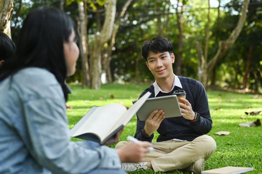Smiling young asian male checking university schedule on digital tablet and talking with his friend while sitting on green grass in campus.