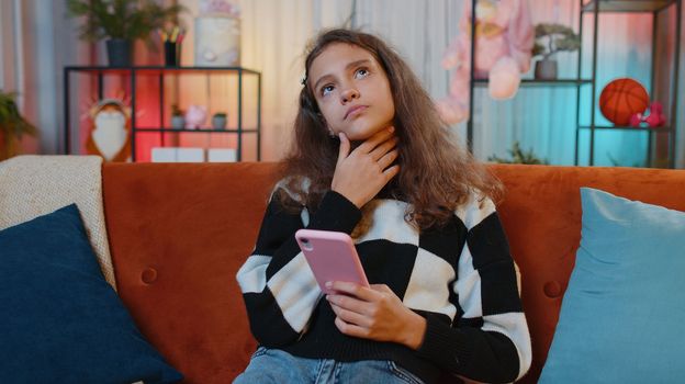 Preteen girl texting share messages content on smartphone social media applications online watching relax movie, news. Young child, kid at home in night evening living play room sitting on orange sofa