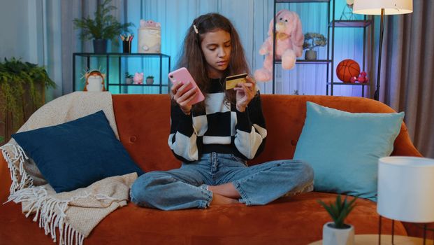 Preteen girl using credit bank card and smartphone while transferring money, purchases online shopping, order food delivery at home apartment indoors. Young teenager child, kid in playroom on couch