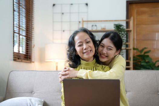 Happy adult granddaughter and senior grandmother having fun enjoying talk sit on sofa in modern living room, smiling old mother hugging young grown daughter bonding chatting relaxing at home together.