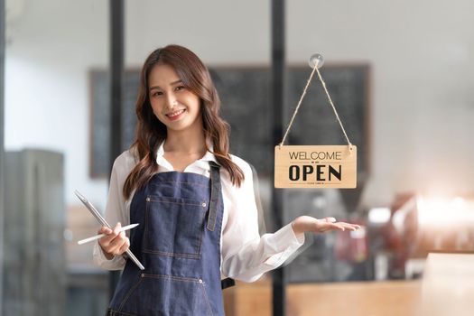Beautiful asian young barista woman in apron holding tablet and standing in front of the door of cafe with open sign board. Business owner startup concept...