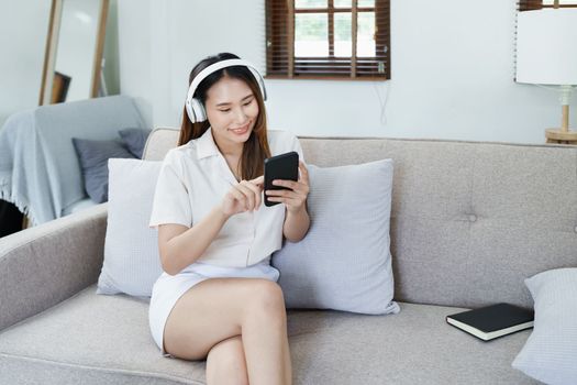 Portrait of asian woman using smart phone mobile and headphones relaxing on sofa at home.