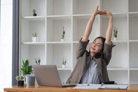Asian businesswoman stretches her arms to relax her tired muscles from working at her desk all day at the office...