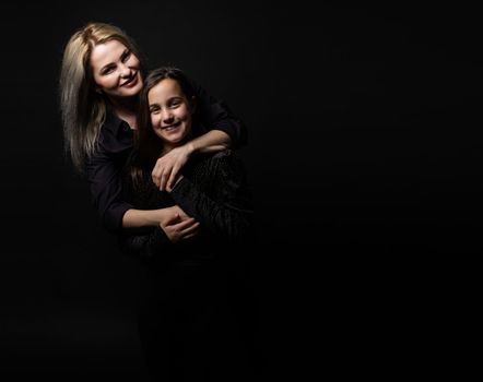 Young mother and her little daughter are looking at camera and smiling, sitting against black wall.