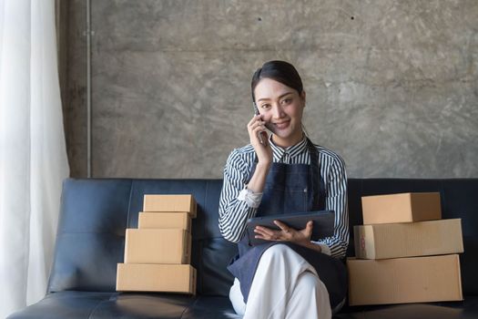 Startup small business entrepreneur SME freelance woman work with smart phone, Young Asian small business owner use computer, online market packing box delivery, SME e-commerce telemarketing concept..