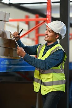 Smiling asian male worker using digital tablet and checking quantity of storage product on shelves full of packed boxes.