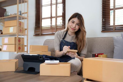Asian business woman leaning on the sofa use laptop computer checking customer order online shipping boxes at home. Starting Small business entrepreneur SME