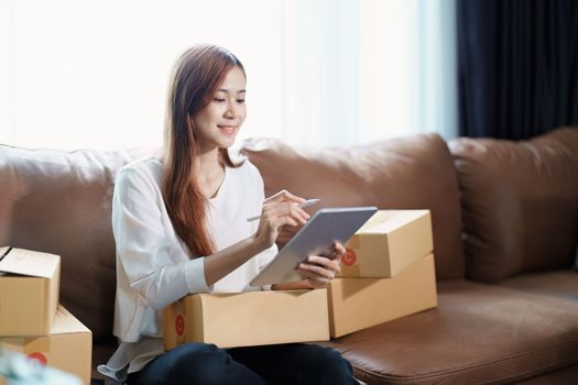 Starting small business entrepreneur of independent young Asian woman online seller is using tablet computer and taking orders to pack products for delivery to customers. SME delivery concept.