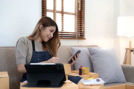 Startup SME small business entrepreneur woman using phone call receive from customer checking product on stock at home office, online marketing packaging delivery box