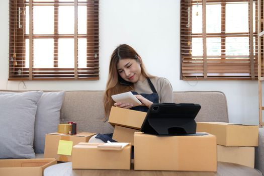 Startup SME small business entrepreneur woman using phone call receive from customer checking product on stock at home office, online marketing packaging delivery box