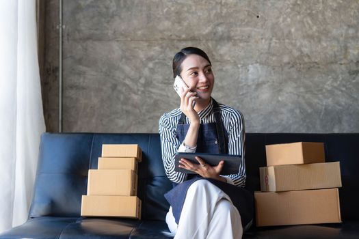 Startup small business entrepreneur SME freelance woman work with smart phone, Young Asian small business owner use computer, online market packing box delivery, SME e-commerce telemarketing concept..