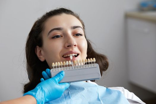 Dentist choosing color of tooth enamel for patient. Dentist applying sample from tooth enamel scale to caucasian female patient teeth