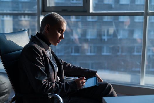 Business woman with short hair sits by the window and writes on a digital tablet with a stylus pen.