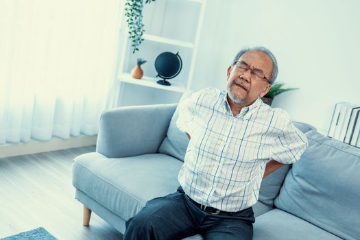 An agonizing senior man in need of assistance while sitting on his sofa at home, suffering from back pain. Senior care, nursing home for pensioners, deteriorating health of old age.