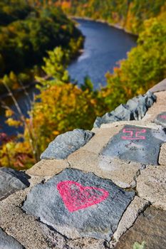 Image of Detail stone wall with pink heart chalk art and soft river with fall forest behind