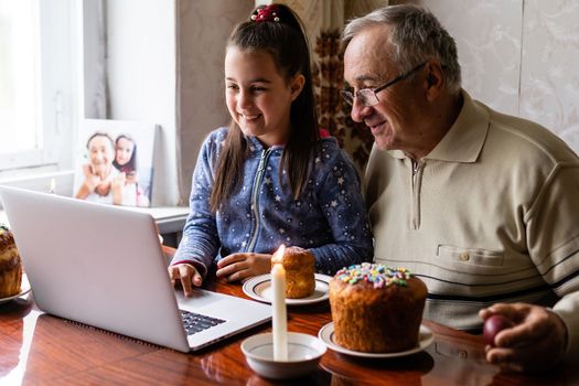 grandfather and granddaughter are talking via video link to their friends. Decorated table with colorful eggs and cake. Chatting during the COVID pandemic and the Easter holidays.