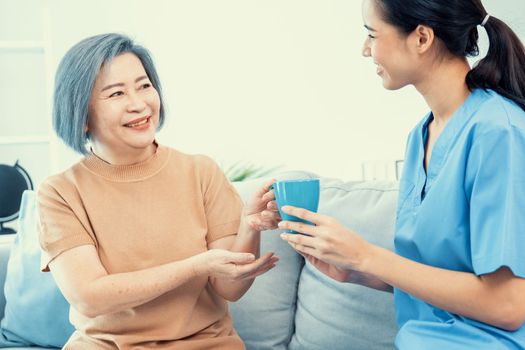 Female care taker serving her contented senior patient with a cup of coffee at home, smiling to each other. Medical care for pensioners, Home health care service.