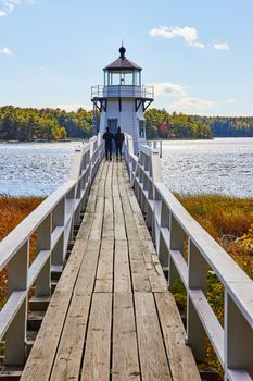 Image of Wood bridge leads to small white Maine lighthouse with two tourists walking