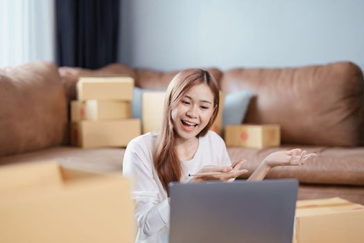 Starting small business entrepreneur of independent Asian female online seller talking on computer to video with a customer and packing products for delivery to the customer. SME delivery concept.