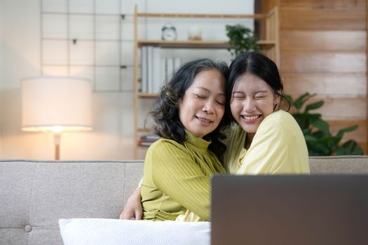 Happy adult granddaughter and senior grandmother having fun enjoying talk sit on sofa in modern living room, smiling old mother hugging young grown daughter bonding chatting relaxing at home together.