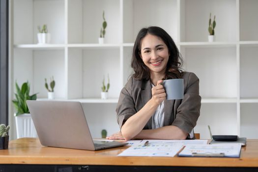 Beautiful young Asian businesswoman smiling holding a coffee mug and laptop working at the office...
