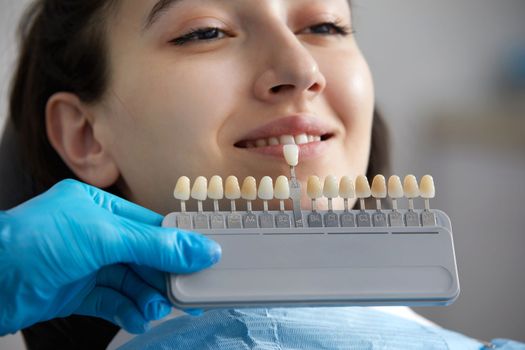 Dentist choosing color of tooth enamel for patient. Dentist applying sample from tooth enamel scale to caucasian female patient teeth