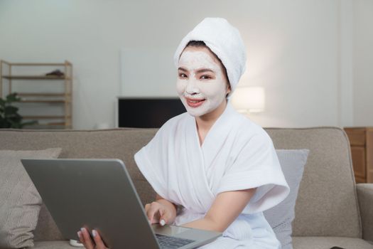 Woman with facial mask using laptop at home. Skin care, face treatment, spa, cosmetology and natural beauty concept.
