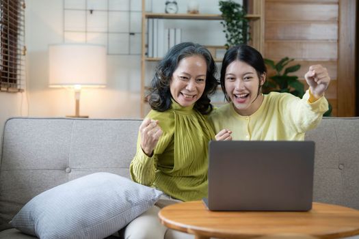 Happy adult granddaughter and senior grandmother having fun enjoying talk sit on sofa in modern living room, smiling old mother hugging young grown daughter bonding chatting relaxing at home together.