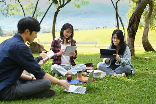 Group of university students working and reading book while sitting together on green lawn at university campus.