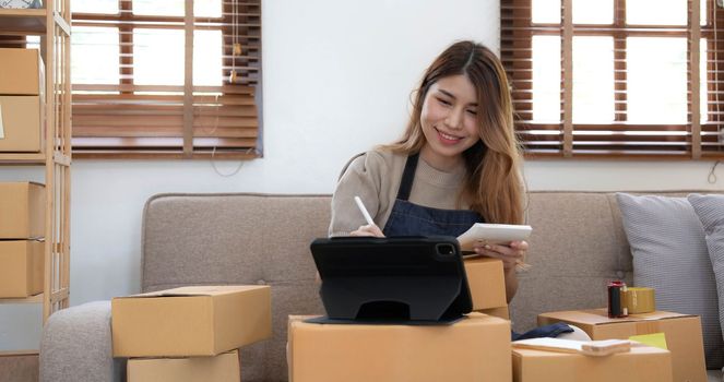 Asian business woman leaning on the sofa use laptop computer checking customer order online shipping boxes at home. Starting Small business entrepreneur SME