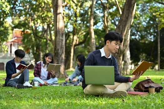 Asian man student reading book on green lawn at university campus. Youth lifestyle, university and friendship concept.