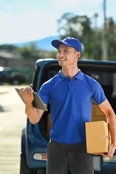 Portrait of delivery man wearing blue uniform with cardboard in hands, searching the address on digital tablet.