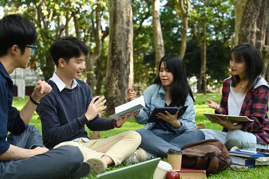 Group of college students spending time together reading book, working on group project and communicating while sitting in the park. 