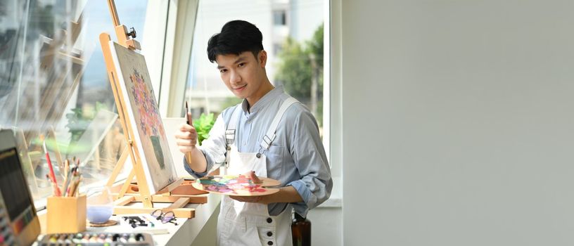 Asian man artist holding palette and paint brush standing in bright art studio. Panoramic web banner with copy space on the right. 