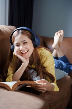 Asian woman reading book and wearing headphones relaxing on sofa at home.
