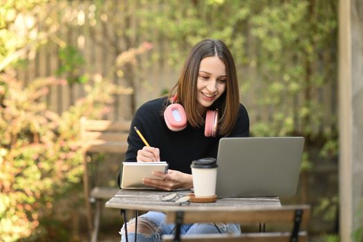 Smiling caucasian woman looking at laptop screen and writing down in notebook while sitting at outdoor.